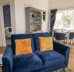 A living area with a blue sofa with two orange cushions a dining table with four chairs and a grey cabinet at Primrose End in Jacobstow