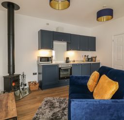 A kitchen area with navy cabinets and appliances next to a wood burning stove and a blue sofa with yellow cushions at Primrose End in Jacobstow