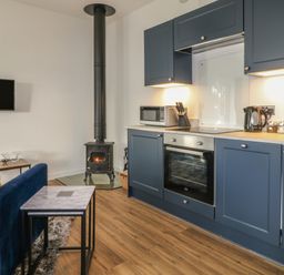 A kitchen with blue cabinets a built in oven microwave kettle a wood burning stove and a blue sofa with nested tables at Primrose End in Jacobstow