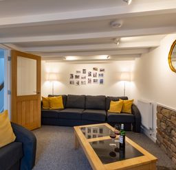 A living room with a dark sofa and yellow cushions two floor lamps a coffee table with wine glasses and a bottle a wooden door and a round mirror on the wall at Rose Cottage in St Ives