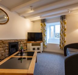 A living room with a stone wall fireplace a TV on a white cabinet a gray sofa with yellow cushions a wooden coffee table with two wine glasses and a bottle a window with floral curtains and a floor lamp at Rose Cottage in St Ives