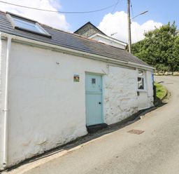 A small white cottage with a blue door and window on a sloping road at The Old Stable in Porthallow