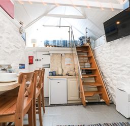 A small kitchen area with a wooden staircase leading to a loft bed and dining table with chairs in a white stone walled room at The Old Stable in Porthallow