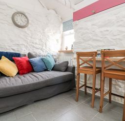 A small living area with a grey sofa with colorful cushions a wall clock wooden chairs and a narrow wooden table at The Old Stable in Porthallow