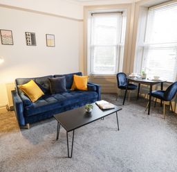 A living room with a blue sofa yellow and blue cushions a black coffee table and a small dining table with two chairs at Sapphire Suite in Barmouth