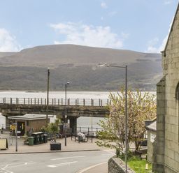 A street with a stone building by a waterfront with a bridge and hills in the background at Rose Suite in Barmouth