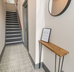 A hallway with patterned tile floor a wooden table with a framed sign a round mirror and stairs leading up at Rose Suite in Barmouth