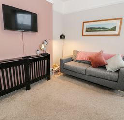A living room with a wall mounted television above a black radiator cover and a gray sofa with cushions and a blanket at Rose Suite in Barmouth