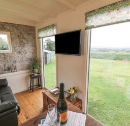 A sitting area with a black sofa a television on the wall and large windows showing a grassy field at Shepherds Hut in Colton near Rugeley Staffordshire
