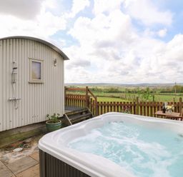 An outdoor hot tub near a small shed with an outdoor shower and a wooden table with chairs at Shepherds Hut Colton near Rugeley Staffordshire