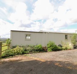An exterior view of a corrugated metal building with two windows surrounded by plants and a gravel driveway at Shepherds Hut in Colton near Rugeley Staffordshire