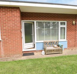 A brick exterior with a glass door next to a window and a wooden bench on a concrete path with grass in front at The Nook in Mundesley