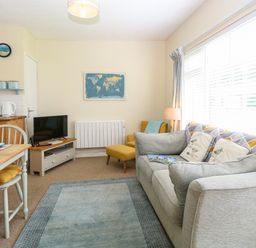 A living room with a gray sofa and cushions a small TV on a stand a yellow armchair a wooden dining table with chairs and cushions a world map wall art and large windows with blinds at The Nook in Mundesley