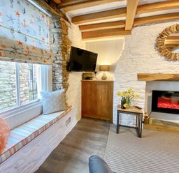 A sitting area with a window seat cushions a tv on a wooden cabinet a table with flowers and a fireplace in Pilchard Cottage in Brixham