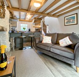 A living room with a gray sofa and a kitchen area with bar stools and wooden beams at Pilchard Cottage in Brixham