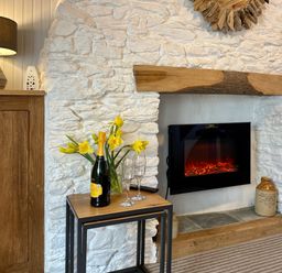 A fireplace with a wooden mantel and electric fire with a small table holding a bottle of prosecco and two glasses next to a cabinet with a lamp and yellow flowers at Pilchard Cottage in Brixham