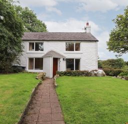 A white two story house with a tiled roof and four windows with a paved path leading to the entrance surrounded by grass and trees at Penllain in Newport Pembrokeshire
