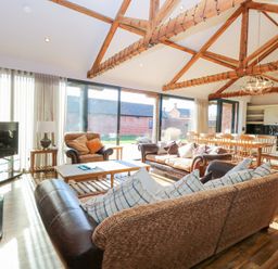 An open living room and kitchen with wooden beams on the ceiling large windows sofas armchair coffee table dining table and chairs a television and kitchen cabinets at Shorthorn Barn Attlebridge near Taverham