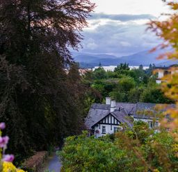 A house surrounded by trees and bushes with mountains and a lake in the background at Fellside Lodge in Bowness-On-Windermere