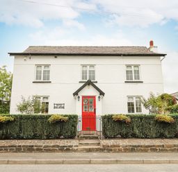 A white two-story house with a red door and black fence surrounded by greenery at The Old Post Office in Llandrindod Wells