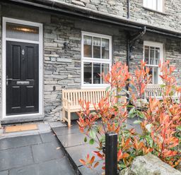 The front entrance of a stone house with a black door two wooden benches and a plant with orange leaves at Netherbeck in Ambleside