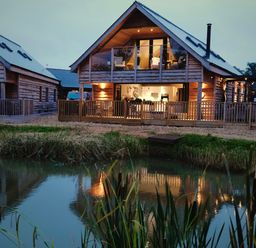 A wooden two-story house with a balcony and patio lights next to a pond at Lake View Lodge in Thorpe-On-The-Hill