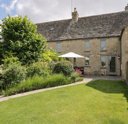 A stone house with a garden lawn and green bushes with patio furniture and an umbrella at Little Owl Barn in Burford