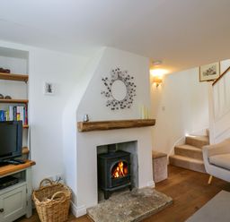 A living room with a lit stove fireplace a TV on a stand shelves with books a basket stairs and a chair at Little Owl Barn in Burford