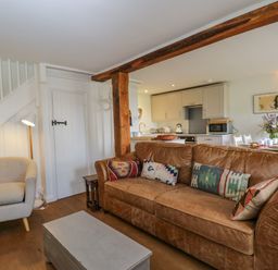 A living room with a brown leather sofa and patterned cushions a beige armchair a gray coffee table and a kitchen with dining table and flowers at Little Owl Barn in Burford