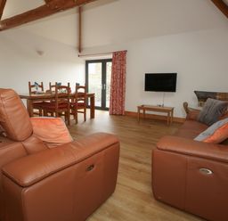 A living room with brown leather sofas a wooden dining table with chairs a wall-mounted TV and wooden beams at Swallows Nest in Puxey near Sturminster Newton