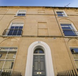 The front facade of a three-story building with a grey door numbered 23 and windows with black railings at 23 Daniel Street in Bath