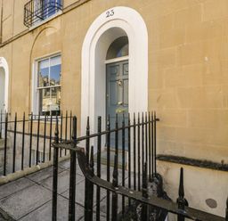 An exterior view of a building entrance with a blue door numbered 23 and black metal railings at 23 Daniel Street in Bath