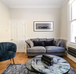 A living room with a gray sofa black pillows a blue chair round tables with a leaf design and a black and white car picture on the wall at 23 Daniel Street in Bath