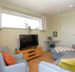 A living room with a gray armchair and sofa a TV on a wooden stand and a window with patterned curtains at The Wren in Wem