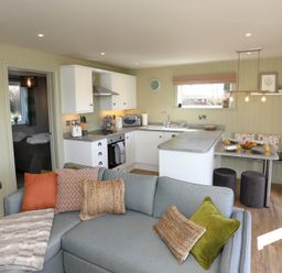 An open living space with a grey sofa and cushions next to a white kitchen with a dining nook at The Wren in Wem