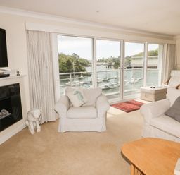 A living room with white armchairs and a sofa facing a glass door with a view of boats on water at Creekside House in Fowey