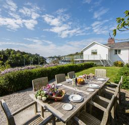 An outdoor dining table with plates and drinks in a garden with a white house and water view at Creekside House in Fowey