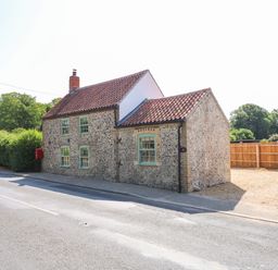 A traditional stone cottage with green windows by a road and a gravel driveway at Postbox Cottage in Feltwell
