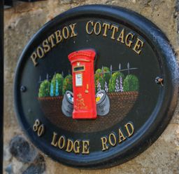 A decorative black circular plaque with a red postbox and garden scene showing Postbox Cottage 80 Lodge Road
