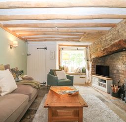 A living room with wooden beams on ceiling a stone fireplace with a wood stove a sofa an armchair and a window seat at Tuesday Cottage in Bourton-On-The-Water