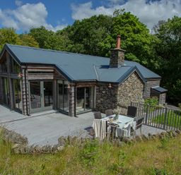 A stone and wood house with a metal roof and outdoor dining table on a patio surrounded by trees at The Owl's Shack in Kendal