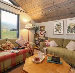 A living room with two sofas cushions and blankets a wooden coffee table with books and cups a floor lamp and a window showing a grassy landscape at The Badger's Shack in Garnett Bridge Longsleddale Near Kendal