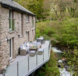 An outdoor deck with seating and dining area next to a stone house overlooking a stream with trees at The Old Water Mill in Kendal