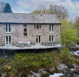 A stone house with a slate roof and a balcony overlooking a river and rocks at The Old Water Mill in Kendal