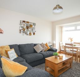 A living room with a dark sofa and cushions a wooden coffee table and a dining table with chairs near a window at Sandy Toes in Rhosneigr