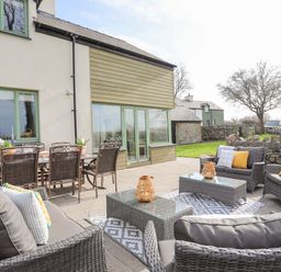 An outdoor patio with wicker seating and glass tables next to a house with green window frames at Dolwaenydd in Brynsiencyn