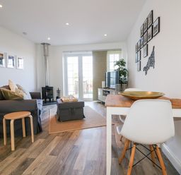 A living room with a gray couch, wooden stool, coffee table, dining table with white chairs, TV, and wood-burning stove at Badgers Way in Tintagel
