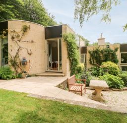 An exterior view of a modern house with a patio garden bench and table surrounded by plants and trees at Under Catswood in Elcombe near Stroud