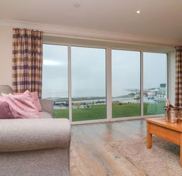 A living room with a gray sofa with pink pillows a wooden coffee table with a champagne bucket and glasses and large windows with a view of a grassy area and coastline at The Lobster Pot in Westward Ho