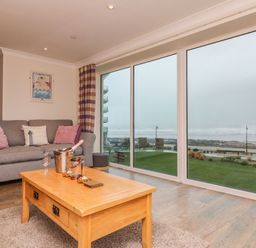 A living room with a gray sofa with pink cushions and a wooden coffee table next to large windows overlooking the sea at The Lobster Pot in Westward Ho